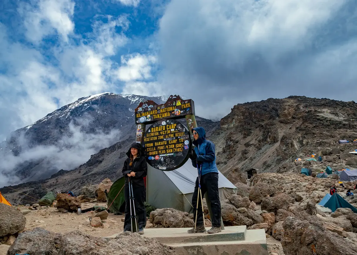 Two climbers at the Barafu Camp sign with tents on the rocky ridge and Kibo's snow behind