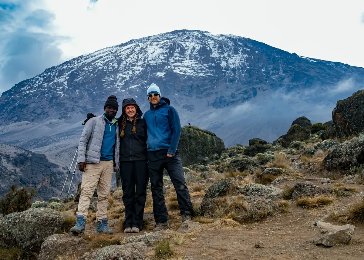 A honeymoon couple and their guide smiling in the alpine zone, with Kibo's snow-covered south face behind them