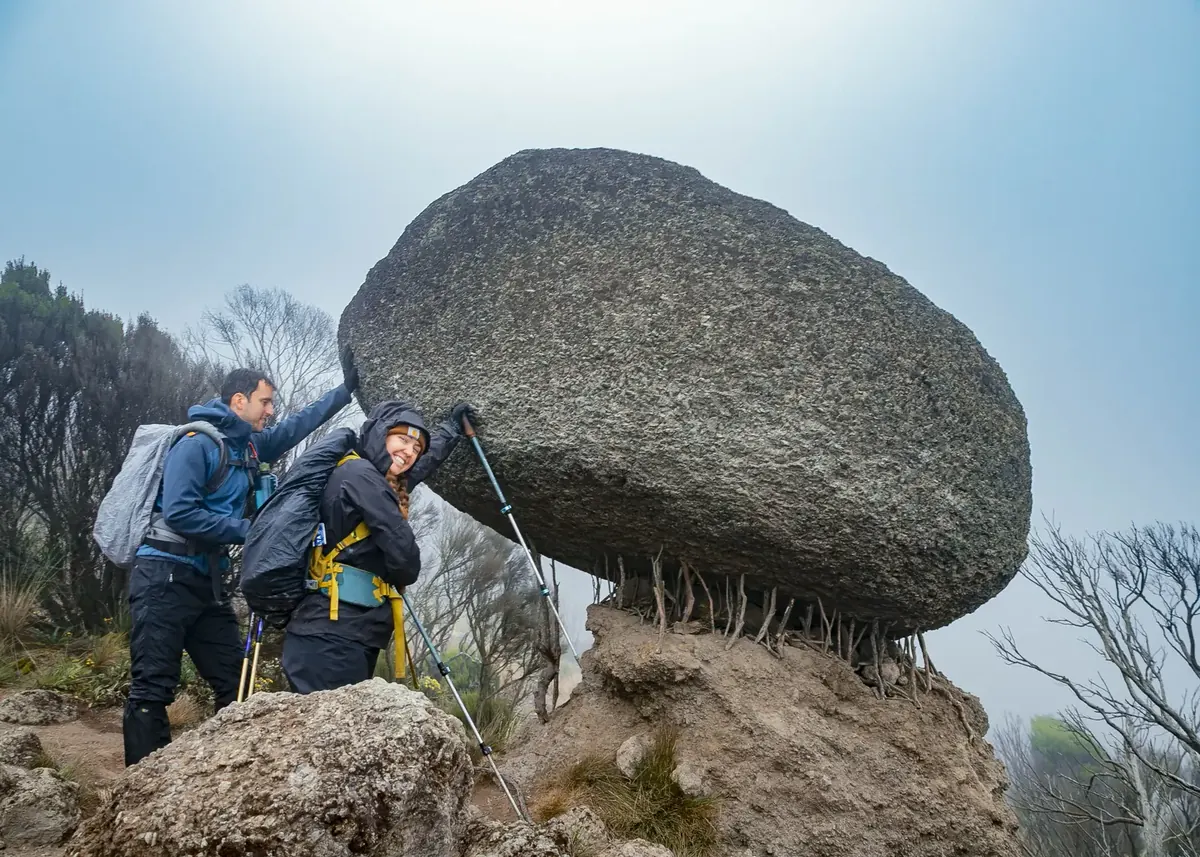 A honeymoon couple in rain gear laughing as they pretend to hold up a huge balancing boulder in the mist on Kilimanjaro