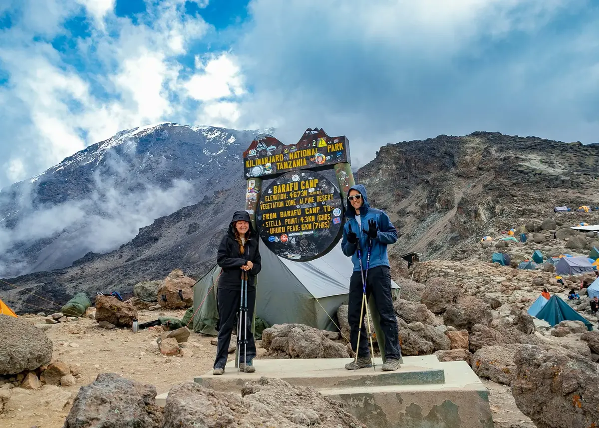 A couple in warm jackets at the Barafu Camp sign with tents on the rocks and the snow of Kibo behind