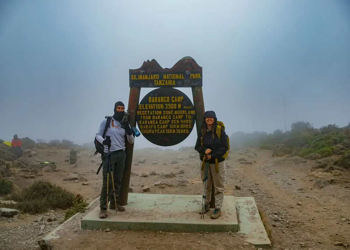 A couple in warm layers at the Barranco Camp sign, 3,900 m, in cloud on Kilimanjaro