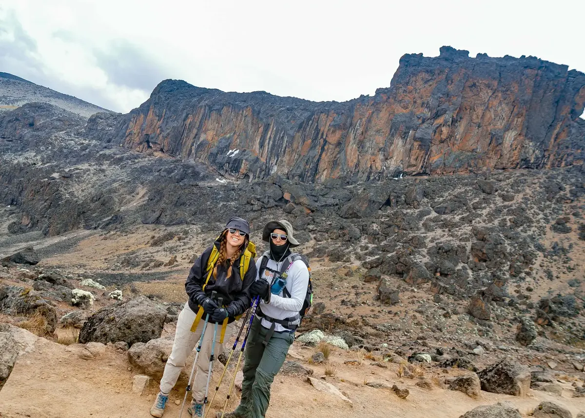 A honeymoon couple in sunglasses and sun hats on the trail below tall volcanic cliffs in Kilimanjaro's alpine desert