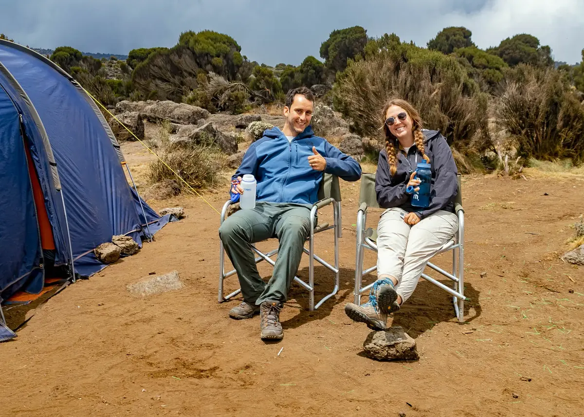A honeymoon couple relaxing in folding camp chairs beside their blue tent among the heath on Kilimanjaro