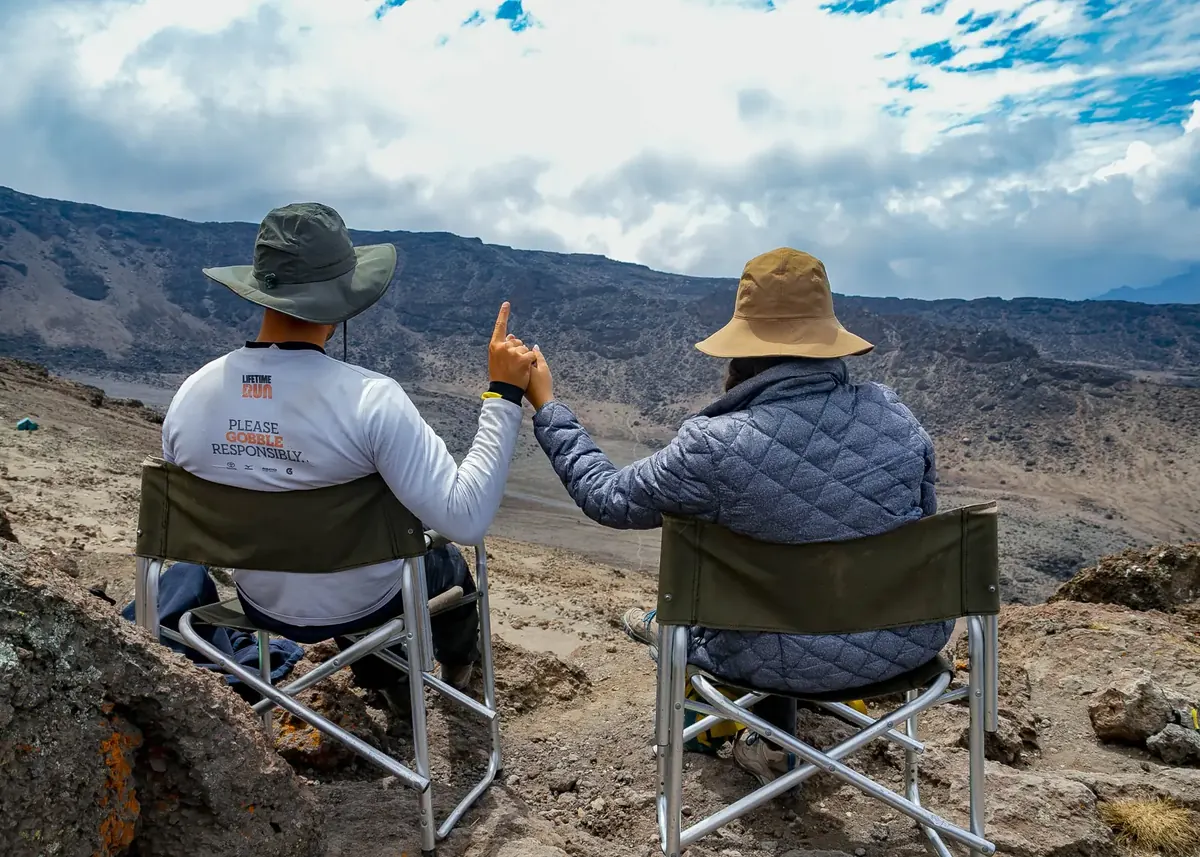 A couple in camp chairs high on Kilimanjaro, the woman pointing to the view over the rocky alpine desert