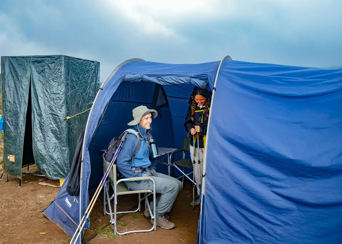 A couple in their blue mess tent with folding chairs and a table, next to a green private toilet tent, at camp on Kilimanjaro