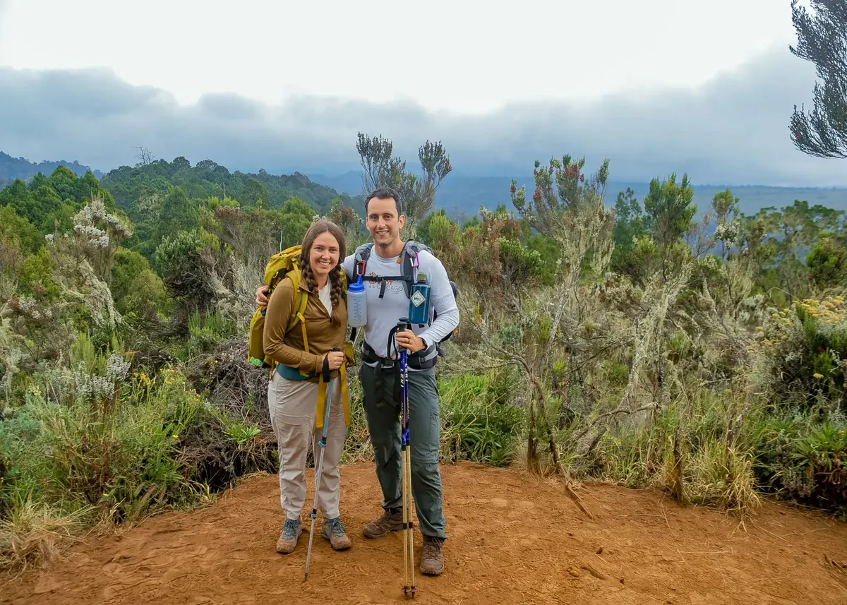 A honeymoon couple with trekking poles among giant heather and lichen-hung trees, with forest and clouds below