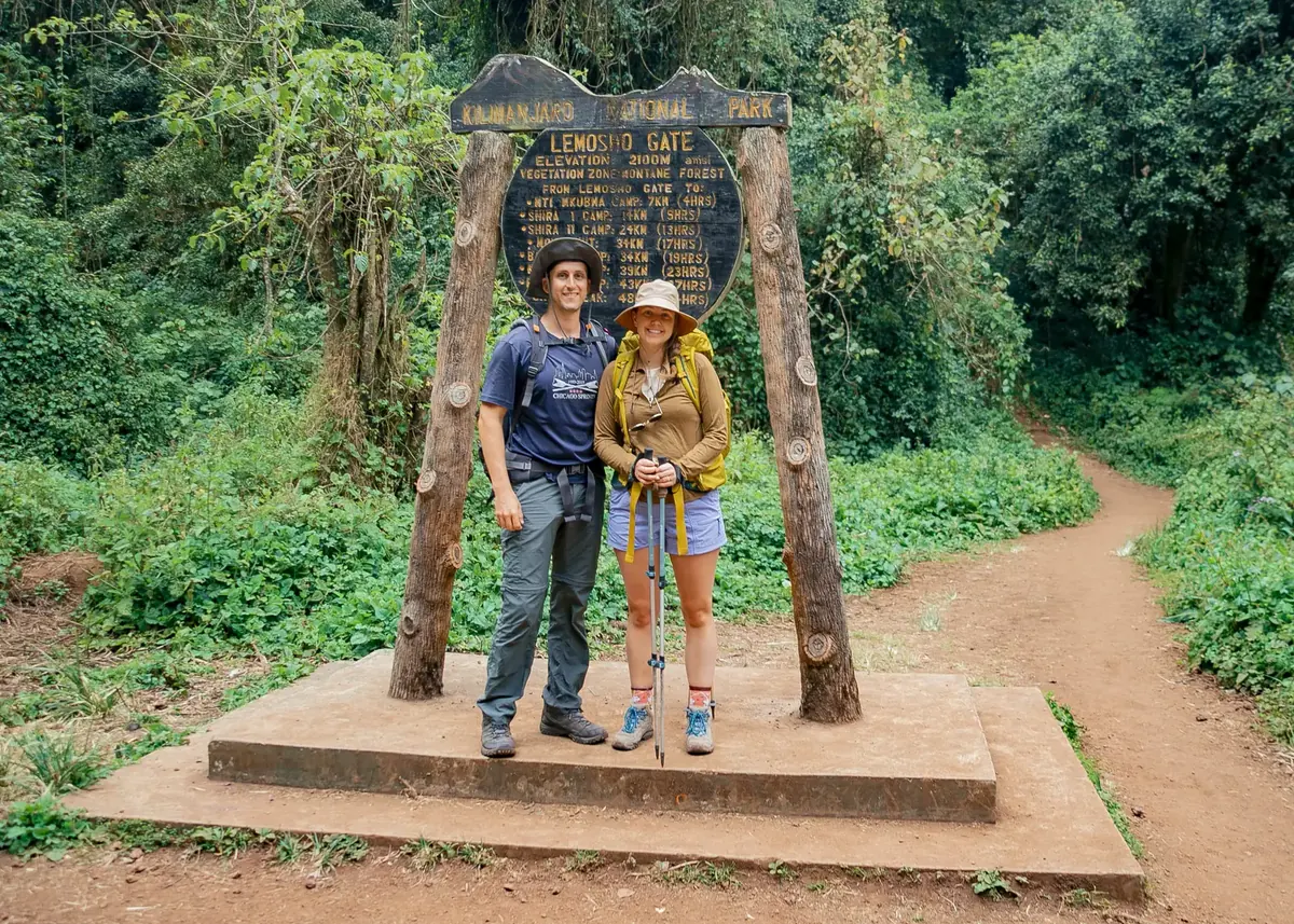 A honeymoon couple with trekking poles at the Lemosho Gate sign of Kilimanjaro National Park, at the start of the forest trail