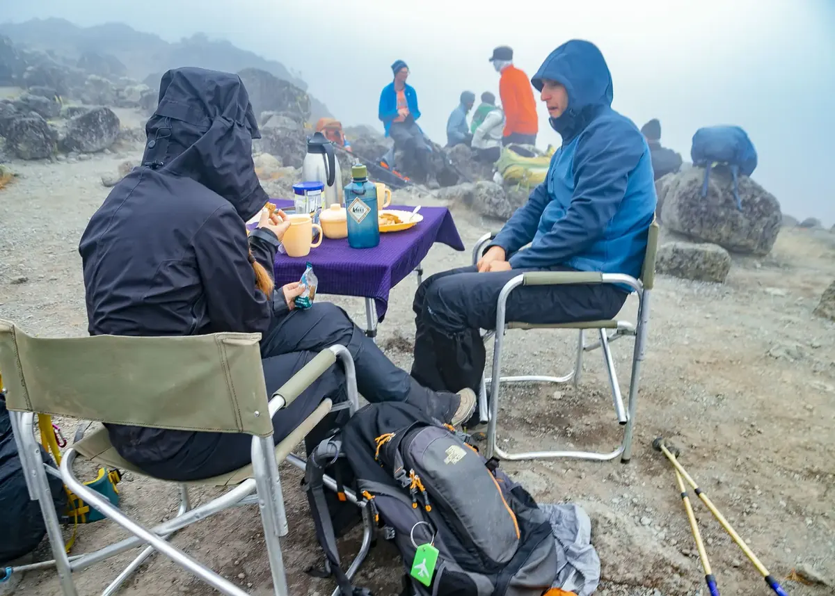 A couple in hooded jackets at a lunch table with a flask and mugs in the mist on Kilimanjaro