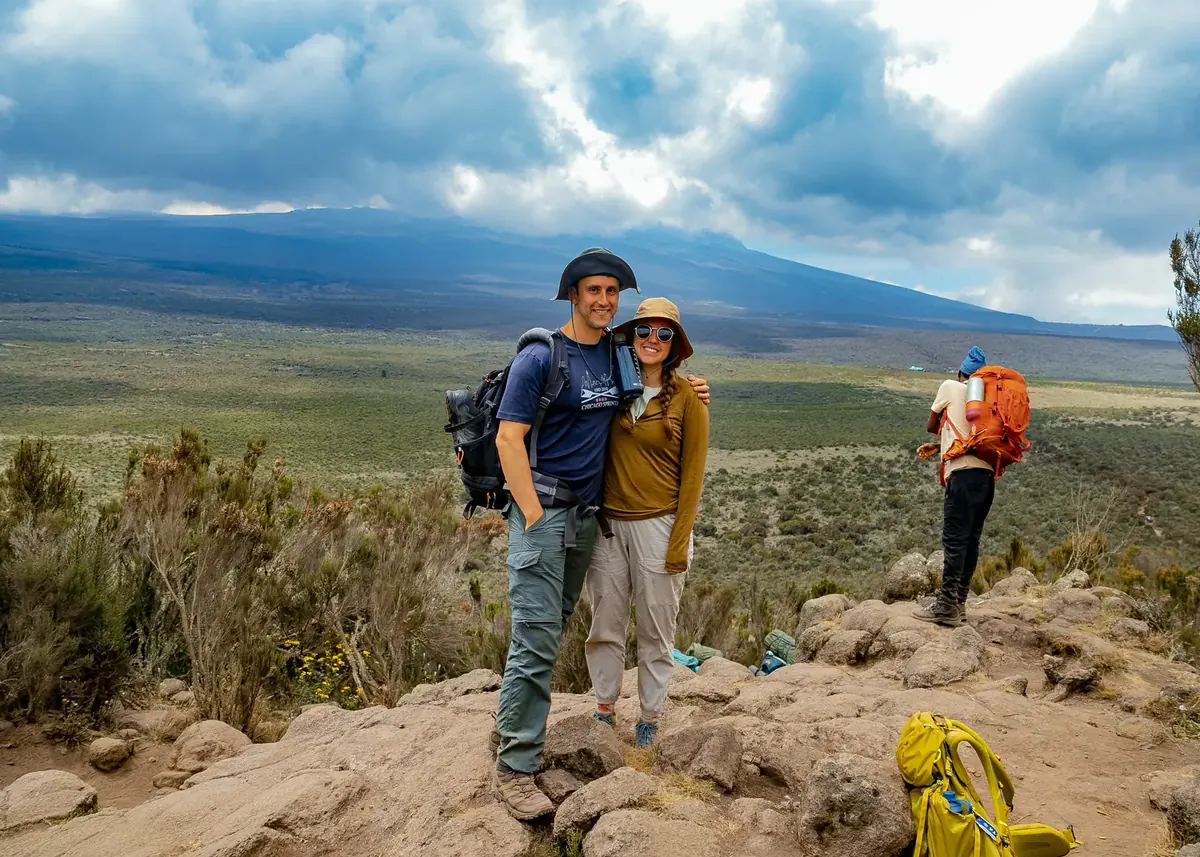 A couple in sun hats on a rocky lookout high above the plains of Kilimanjaro, with a porter carrying a pack nearby