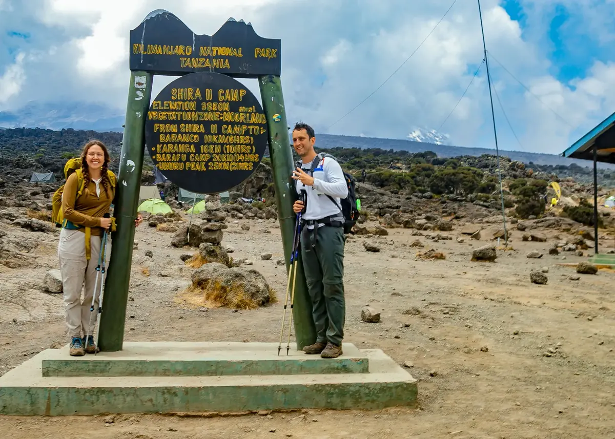 A smiling couple with trekking poles at the Shira 2 Camp sign, 3,850 m, with tents and Kibo's snow behind