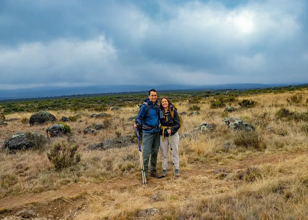 A couple in rain jackets on the moorland of Kilimanjaro, with lichen-covered rocks and clouds over the plains