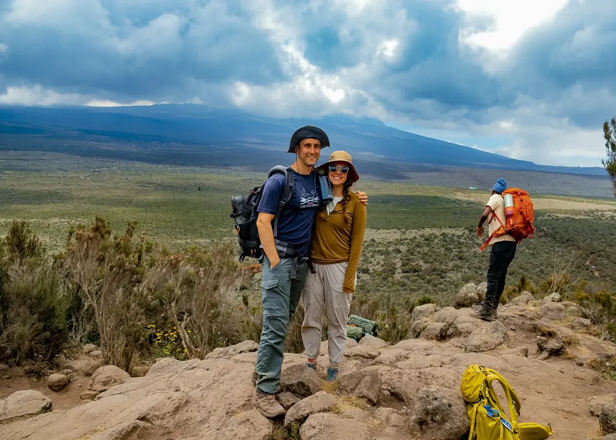 Two climbers on a rocky lookout above the wide Shira Plateau, with Kibo under the clouds and a porter on the trail