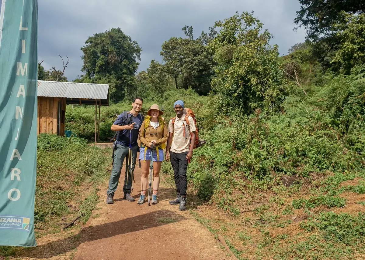 A honeymoon couple with trekking poles and their guide with a large orange pack on the trail beside a Kilimanjaro banner