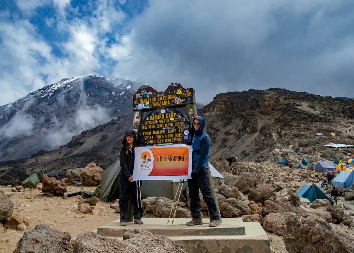A just-married couple in rain jackets holding a Jaynevy Tours Just Married banner at the Barafu Camp sign, with Kibo's snow and the tents behind