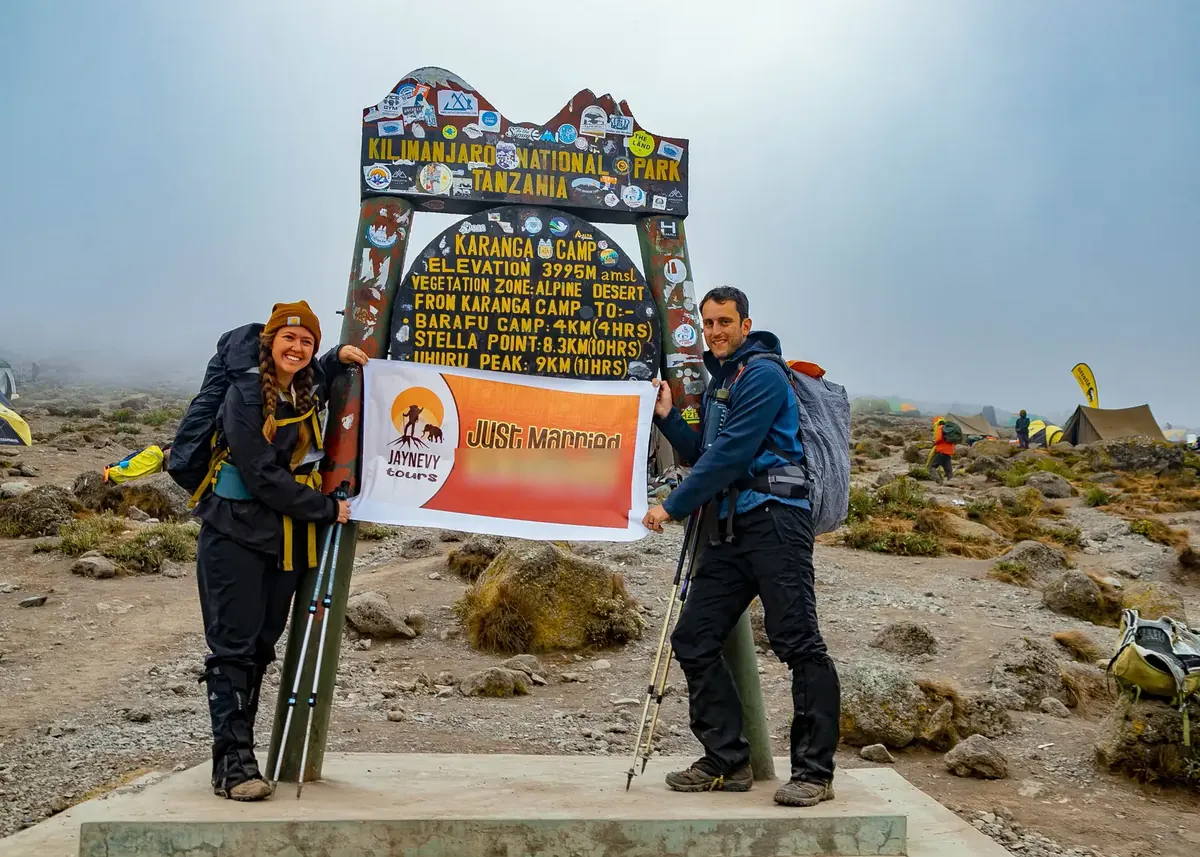 A just-married couple holding a Jaynevy Tours Just Married banner at a camp sign on Kilimanjaro