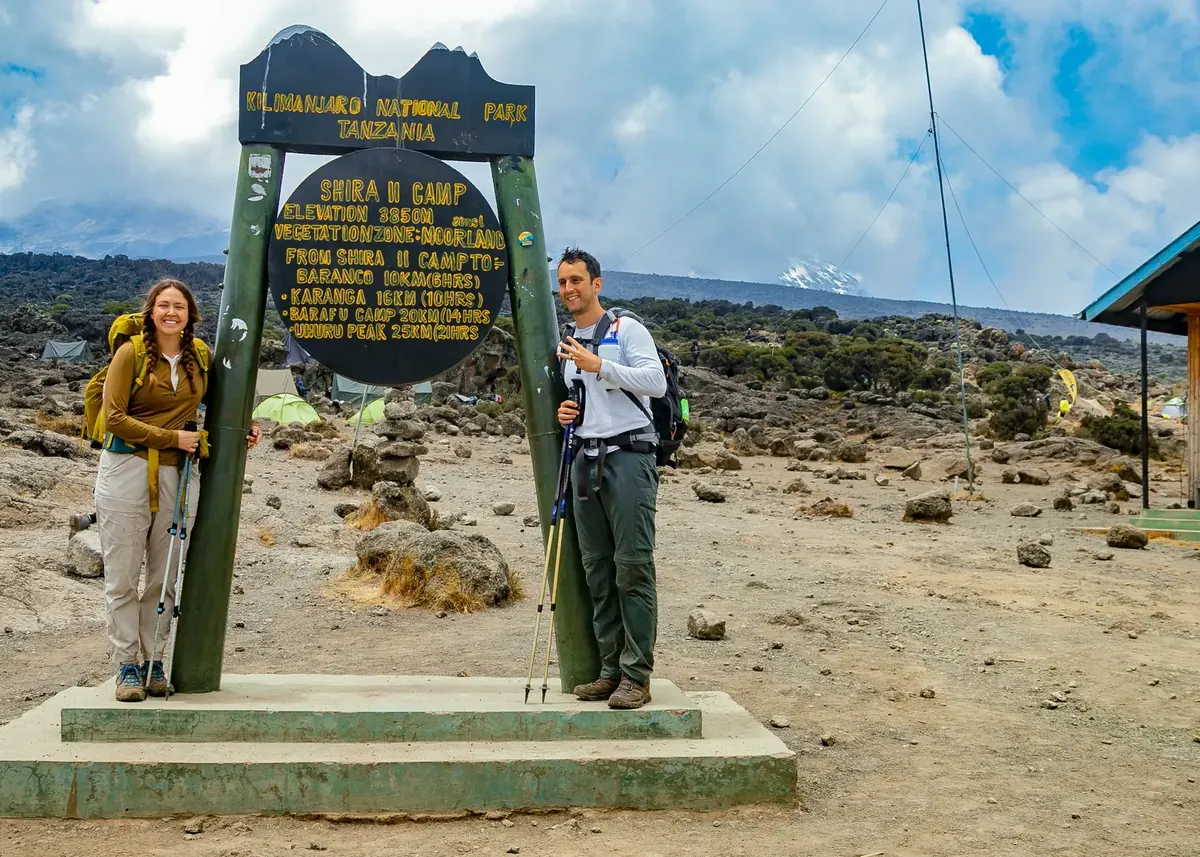 Two climbers at the Shira 2 Camp sign, 3,850 m, with the moorland and Kibo's snow behind